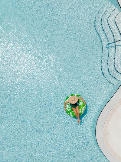 Freeform pool with a women floating in a green ring. She is wearing a black swimsuit and a wide rim straw hat. 