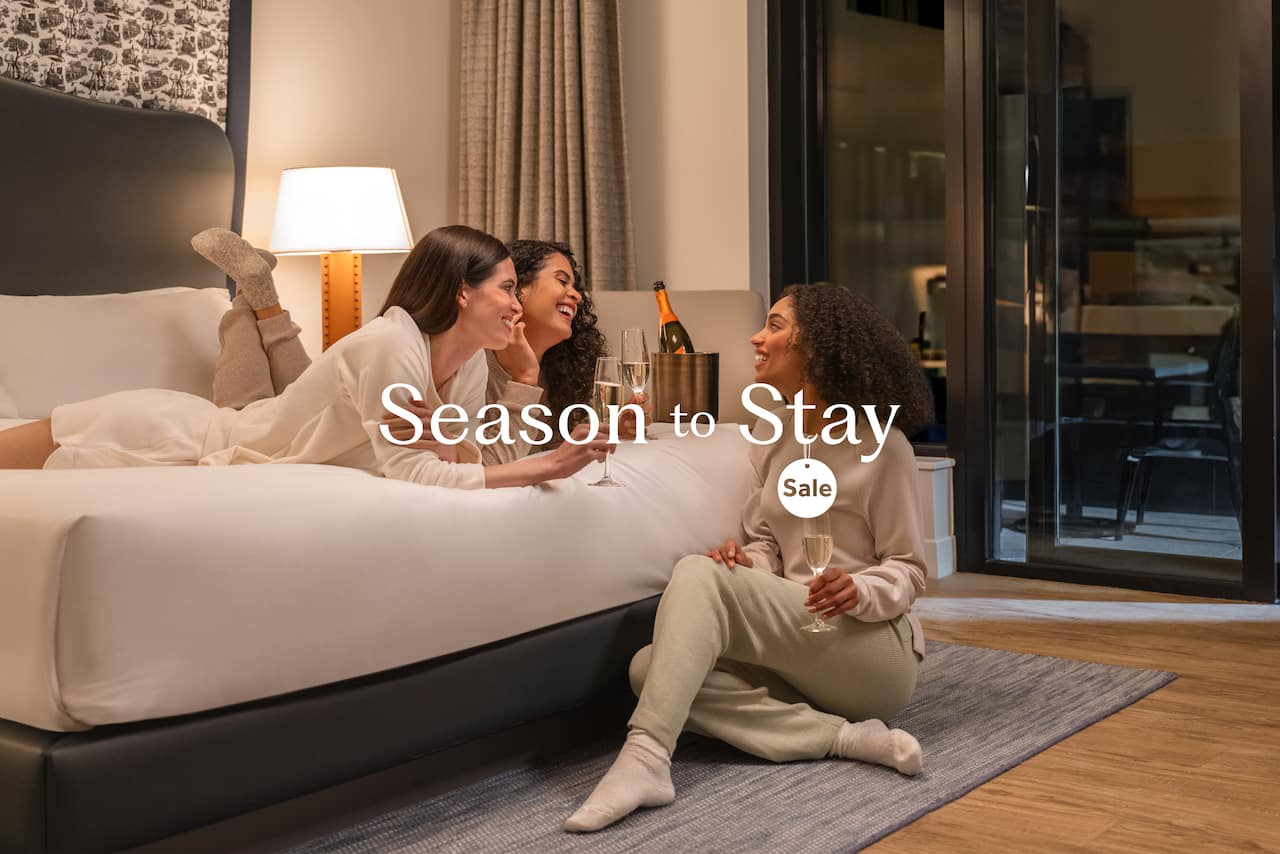 Three women enjoying a cozy evening on a hotel bed, laughing and chatting, with champagne glasses in hand, under warm lighting; text overlay reads "Season to Stay Sale."