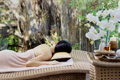 A lady lying on a message table is covered by a towel with her back to the camera. There are massage oils and flowers in the foreground and a tranquil garden in the background. 
